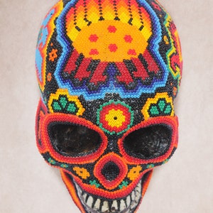 May include: A colorful beaded skull with a black background and intricate designs in shades of red, orange, yellow, green, and blue. The skull is decorated with flowers and other patterns.