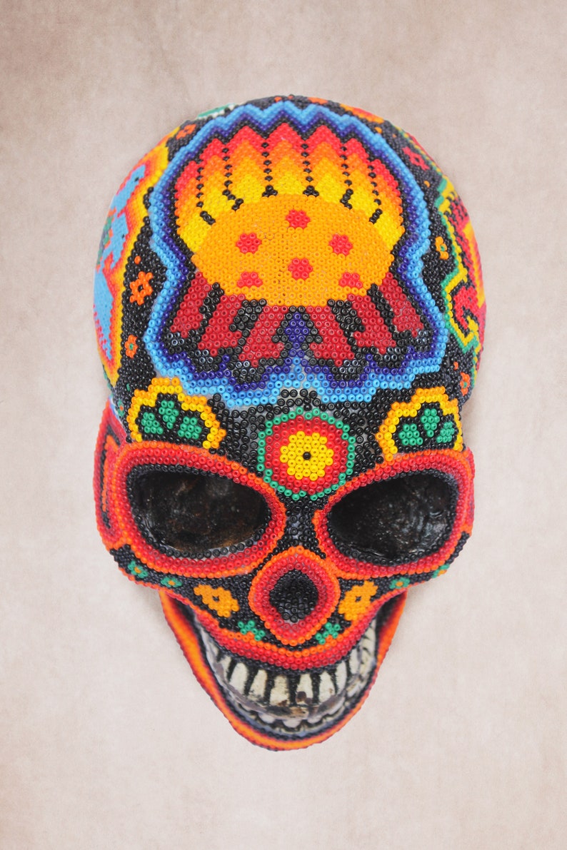 May include: A colorful beaded skull with a black background and intricate designs in shades of red, orange, yellow, green, and blue. The skull is decorated with flowers and other patterns.