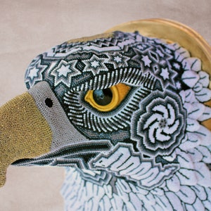 May include: A beaded eagle head sculpture with a gold beak and a detailed eye. The eagle's feathers are white and gray with intricate geometric patterns.