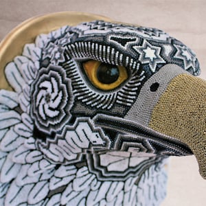 May include: A close-up of a beaded eagle head sculpture. The eagle's eye is yellow, and the beak is gold. The eagle's feathers are beaded in a geometric pattern.