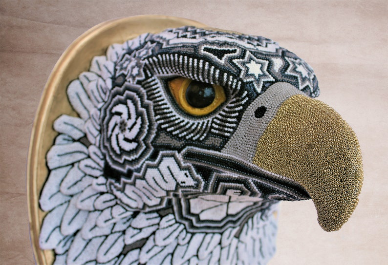 May include: A close-up of a beaded eagle head sculpture. The eagle's eye is yellow, and the beak is gold. The eagle's feathers are beaded in a geometric pattern.