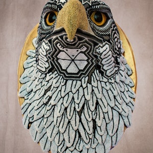 May include: A beaded eagle head sculpture with a gold beak and black and white geometric patterns. The eagle's head is mounted on a gold circular base.