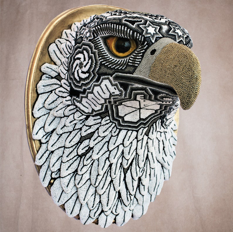 May include: A beaded eagle head sculpture with a gold beak and white and black feathers. The sculpture is mounted on a gold circular frame.