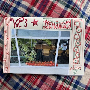 May include: A handmade card with a photograph of a window scene. The card features red and green embroidered text that reads "Vol. 3," "SEEMINGLY," and "MUNDANE photos." The card is decorated with a red star and floral patterned ribbon.