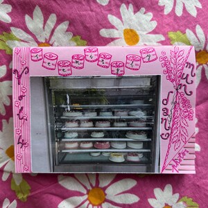 May include: A pink photo frame with a photograph of a glass display case filled with cakes. The frame has pink drawings of cakes and text. The background is a pink floral pattern.