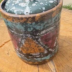 May include: A ceramic jar with a lid, featuring a speckled teal and white pattern on the lid and rim. The jar's body displays a mix of orange, red, and teal hues, with a dark brown base. It sits on a wooden tree stump.