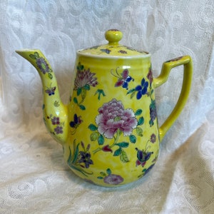 Pode incluir: Uma chaleira amarela com um desenho floral. A chaleira tem uma tampa e um bico. As flores são rosa, azul e verde. A chaleira está sobre uma toalha de renda branca.