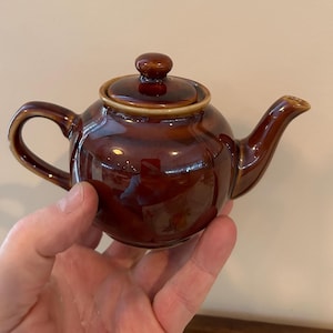 Könnte beinhalten: Eine kleine braune Teekanne aus Keramik mit Deckel. Die Teekanne hat einen runden Korpus und einen Ausguss an der Seite.