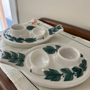 May include: Two white ceramic egg cups with green leaf designs. Each cup has two indentations for holding eggs. The cups have a handle on one side.