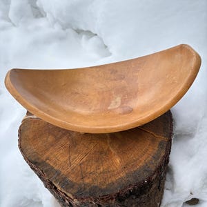 May include: A handcrafted, light brown wooden bowl with a curved, elongated shape. The bowl rests on a section of a tree trunk, showcasing the natural wood grain. The background is a snowy landscape.
