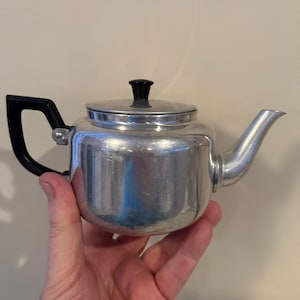 May include: A small silver teapot with a black handle and knob. The teapot has a shiny, metallic finish.