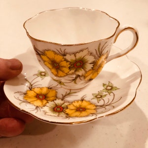 May include: A vintage teacup and saucer set. The white porcelain cup and saucer feature a floral design with yellow and white flowers, green leaves, and gold trim. The cup has a scalloped rim and a curved handle.