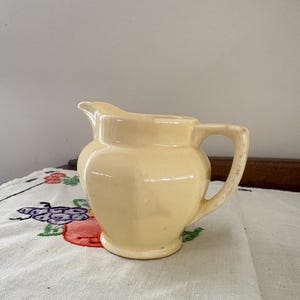 Op de afbeelding: Een kleine, crèmekleurige keramische kan met een handvat en tuit. De kan staat op een wit tafelkleed met geborduurde fruit- en bloemmotieven. De kan heeft een glanzende afwerking.