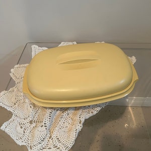 Puede incluir: Un recipiente rectangular amarillo pálido para guardar alimentos con una tapa a juego y un asa central. El recipiente descansa sobre un tapete de ganchillo blanco. Un artículo de cocina vintage. Probablemente de plástico.