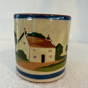 Könnte beinhalten: Ein Keramikbecher mit handgemaltem ländlichem Landschaftsdesign. Das Design zeigt ein weißes Haus mit rotem Dach, grüne Bäume und blaue und braune Streifen um den Sockel. Der Rand ist blau eingefasst.