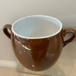 May include: A brown ceramic soup bowl with two handles. The bowl has a glossy finish and a white interior. The handles are curved and attached to the sides of the bowl. The bowl is empty.