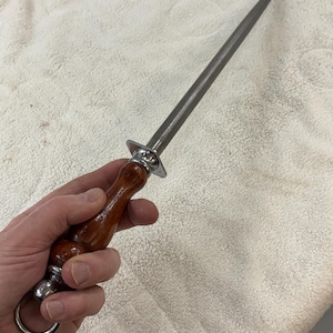 Puede incluir: Una varilla de afilado de cuchillos de acero con un mango de madera. El mango es de color marrón oscuro y tiene un anillo de metal plateado en la base. La varilla de afilado se sostiene en una mano.