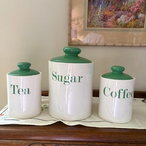 Puede incluir: Tres botes de cerámica blanca con tapas verdes y letras verdes. Los botes están etiquetados como "Té", "Azúcar" y "Café".