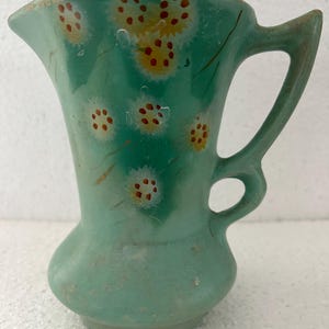 May include: A small, vintage, turquoise-colored pitcher with a handle and a flared spout. The pitcher is decorated with hand-painted floral designs in yellow and red. The pitcher has a bulbous base and a slender neck. The pitcher is likely made of ceramic.