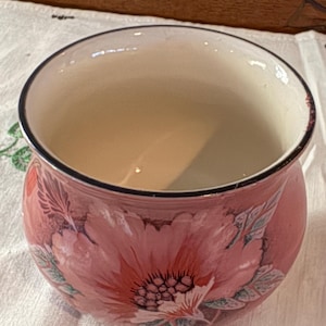 May include: A round ceramic bowl with a pink floral design. The bowl has a cream interior and a black rim. The exterior features a large pink flower with green leaves. The bowl is likely for decorative purposes or serving.
