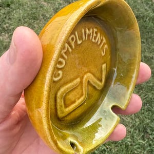 Op de afbeelding: Een vintage keramische asbak in mosterdgeel. De asbak heeft een afgeronde vorm met een kleine inkeping om sigaretten op te leggen. De woorden "COMPLIMENTS" en "CN" zijn aan de binnenkant in reliëf aangebracht. De asbak wordt in een hand gehouden.
