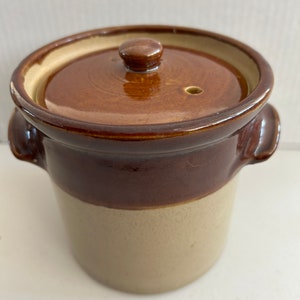 Puede incluir: Una pequeña olla de cerámica redonda con tapa. La olla es de dos tonos, con una parte superior marrón oscuro y una parte inferior beige. Tiene dos asas pequeñas y un pequeño pomo en la tapa.