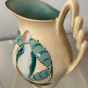 May include: A small, cream-colored ceramic pitcher with a twisted handle and a turquoise interior. The pitcher is decorated with a floral design in shades of turquoise, white, and brown. The pitcher is likely vintage.