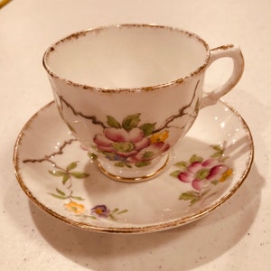 May include: White teacup and saucer set with a floral design. The cup and saucer have a gold trim and feature pink, yellow, and purple flowers with green leaves. The teacup has a handle and sits on the matching saucer.