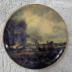 May include: Round decorative plate with a gold rim featuring a landscape painting. The artwork depicts a scene with trees, a river, and figures on horseback, with a cloudy sky in the background.