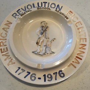 May include: A white ceramic ashtray featuring a relief of a colonial figure. The rim is inscribed with "AMERICAN REVOLUTION BICENTENNIAL" and the dates "1776-1976" in blue, orange, and gold. A commemorative item, likely from the bicentennial.
