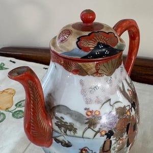 Pode incluir: Uma chaleira de cerâmica vintage com tampa, alça e bico vermelhos. A chaleira é decorada com uma cena de paisagem japonesa em tons de vermelho, dourado e azul. A tampa tem um botão vermelho.