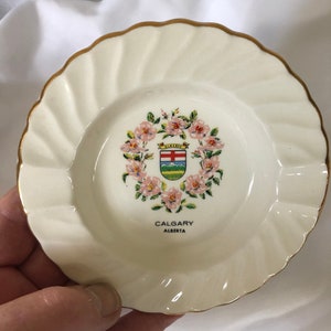 May include: Small, off-white decorative plate with a gold rim and a scalloped edge. The center features a floral wreath surrounding a crest with the words "CALGARY ALBERTA".