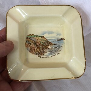 Op de afbeelding: Crèmekleurige vierkante asbak met gouden rand. Het midden toont een schilderachtig beeld van kliffen en de oceaan, met de tekst "Cliffs and Hotel. Land's End." Een decoratief item.