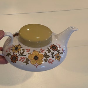 Puede incluir: Una tetera blanca con un patrón floral en tonos amarillos, naranjas y marrones. La tetera tiene una tapa con una parte superior marrón claro.