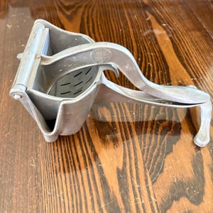 May include: A silver-colored metal citrus press featuring a hinged lever handle and a perforated bowl for extracting juice. The tool has a sturdy construction with a circular embossed logo on the side of the handle and a wide base for stability.