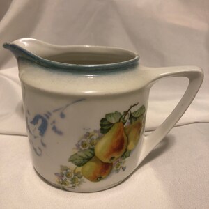 Puede incluir: Jarra de cerámica color crema con asa y boquilla. La jarra presenta un diseño de pera con hojas verdes y peras amarillas. El borde tiene un acento azul y hay un diseño floral azul en el lateral.