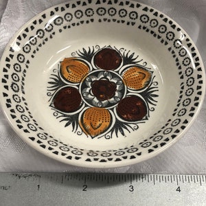 May include: A small, round, cream-coloured ceramic bowl with a decorative floral design. The bowl's interior features a central flower motif with brown, orange, and black accents. The rim is adorned with a repeating pattern of black circles and dots.