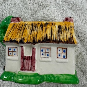 Op de afbeelding: Een keramieken huisje met een rieten dak, rode schoorsteen en groene grasbasis. Het witte huis heeft een rode deur en blauw gestippelde ramen. Aan de linkerkant staat een groene boom.