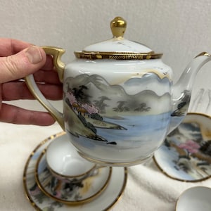 May include: A white porcelain tea set with a landscape design. The set includes a teapot, teacups, and saucers. The teapot and cups have gold accents. The landscape features mountains, water, and trees in shades of blue, grey, and pink.