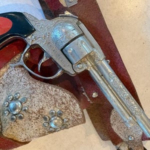 Op de afbeelding: Een speelgoedpistool met een zilveren afwerking, rustend op een bruin leren holster met decoratieve zilveren studs. Het pistool heeft ingewikkelde gravures en een zwarte handgreep met een rode cirkel. De holster heeft een rode rand.