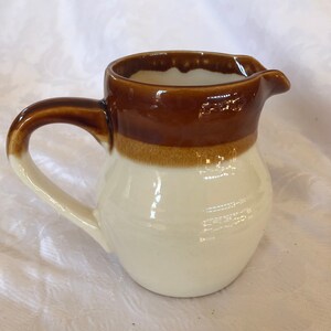 May include: A small, ceramic pitcher with a rounded body and a handle. The top and handle are a glossy brown, with a lighter brown band separating them from the white base. It has a spout for pouring.