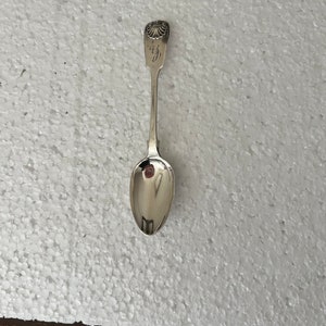 Puede incluir: Una cuchara de plata con un diseño de concha en la parte superior y un cuenco reflectante. La cuchara es probablemente vintage o antigua, con un diseño detallado en el mango. Es un utensilio para comer.