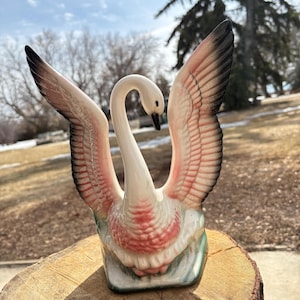 May include: A ceramic swan ornament with open wings, showcasing a white body and neck, pink and black wings, and a black beak. The swan is set on a green and white base, presented on a wooden surface.