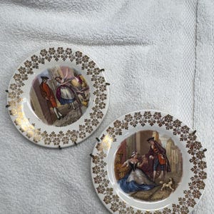 May include: Two decorative porcelain plates with gold floral trim and scenes of people in period clothing. The plates are white with gold trim and are mounted on black metal plate hangers. The plates are on a white, textured surface.