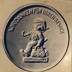 May include: A blue and white ceramic medallion with the text "WIR DANKEN FÜR IHREN BESUCH" encircling a relief of a child carrying a bag and a stick. The child is wearing a hat and seated on a sack. The bottom of the medallion has the text "M.H.H. Keramik".