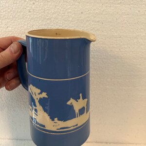 May include: A blue ceramic jug with a cream-coloured rim and handle. The jug features a white relief design of a rider on a horse and a landscape scene. The jug is approximately 18 cm tall.