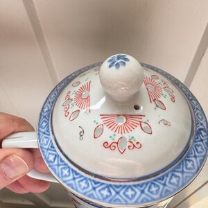 May include: A white ceramic tea cup with a lid, featuring a blue and red floral design. The cup has a handle and a matching lid with a small knob. The rim and handle are trimmed with gold.