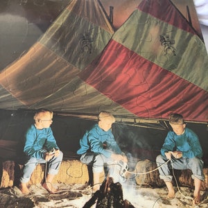 Pode incluir: Um quebra-cabeça vintage com uma cena de fogueira. O quebra-cabeça mostra três crianças com camisas azuis e jeans, reunidas em volta de uma fogueira. Duas tendas com painéis vermelhos, amarelos e verdes estão ao fundo. O texto "Campfire Tales" é visível.