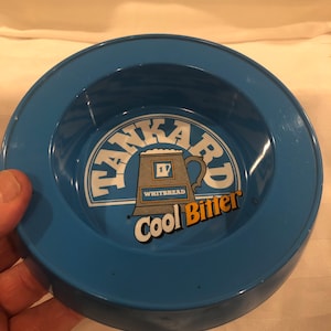 May include: A round, blue ashtray with a central graphic. The graphic features the words "TANKARD" and "Cool Bitter" in white and orange, respectively, with a beer mug illustration. The brand "Whitbread" is also visible.