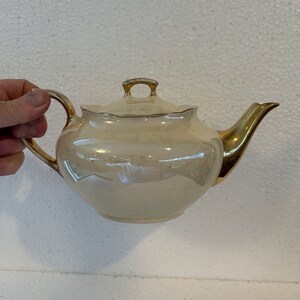 Könnte beinhalten: Cremefarbene Porzellan-Teekanne mit goldfarbenen Verzierungen an Henkel, Ausguss und Deckel. Die Teekanne hat einen runden Körper und einen gewellten Rand am Deckel. Der Griff ist gewölbt und der Ausguss gebogen.